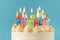 Birthday cake with white cream cheese frosting decorated with multicolored lit happy birthday text shaped candles on a blue