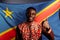 A black African man smiling, posing, showing thumb UP in front of the flag of the Democratic Republic of the Congo.