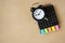 Black analog alarm clock on opened calendar with multi color paper tag on grunge brown paper