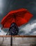 A black backpack protected by a red umbrella against a cloudy sky that threatens rain