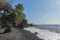 Black beach with lava pebbles and larger stone. Rock cliff of hardened volcanic lava. Slightly wavy sea, foamy water in the surf.