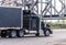 Black big rig classic semi truck with covered semi truck running on the road under old truss bridge