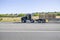 Black big rig semi truck tractor with extended cab transporting lumber cargo on flat bed semi trailer driving on the wide road