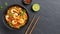 In a black bowl, noodles, veggies, and prawns are stir-fried. slate background. higher perspective