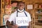 Black business owner wearing a face mask and holding an open sign in front of his shop and smiling