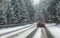 Black car drives on snow covered forest road during snowstorm, trees on both sides, view from car behind. Dangerous driving