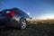 Black car parked at night in green meadows on copy space background of lights of distant city buildings and bright blue sky with