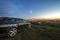 Black car parked at night in green meadows on copy space background of lights of distant city buildings and bright blue sky with