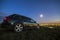 Black car parked at night in green meadows on copy space background of lights of distant city buildings and bright blue sky with