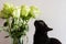 Black cat sniffs flowers in a bouquet of white roses in a glass vase