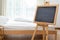 Black chalkboard on easel stand in bedroom with white soft bed background. Interior and decoration concept