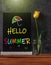 Black chalkboard Hello summer sign mockup, yellow tulip in water glass on dark wooden background. Blackboard with flower