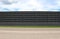 Black concrete surrounding wall with stripe of grass and an asphalt road in front. Cloudy sky above.