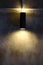A black cylindrical wall lamp that shines up and down, splashes light onto the bare cement wall of the  loft style house