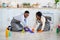 Black guy and his wife in aprons and rubber gloves washing floor in kitchen together, full length portrait