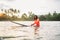 Black long-haired teen man floating on long surfboard, waiting for a wave ready for surfing with palm grove litted sunset rays.