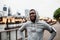 Black man runner with smartphone in an armband on the bridge in a city, resting.