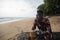 A black man sitting on a tree trunk by the sea enjoys his vacation by enjoying coco juice