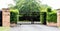 Black metal driveway property entrance gates set in brick fence with garden shrubs and trees in background