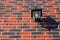 Black metal lantern with dusty glass and energy-saving lamp inside, hanging on wall, casts shadow on the brick facade