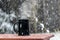 Black mug of hot steaming coffee, on a table outside in the snow. Winter wonderland concept scene.
