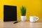 Black notebook with coffee cup and potted plant on wood desk with yellow background.