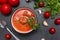 Black plate of red tomato soup with meat meatballs, cilantro on a black background, spoon, cloves and garlic head, fresh cherry ,