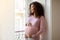 Black pregnant woman gently cradling belly while standing by window at home