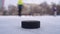 A black puck lies on the ice where men play hockey in the background