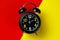 Black round metal alarm clock on legs on a bright composite background of red and yellow. Dial with large numerals.