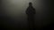 Black silhouette of man standing in smoky dark. Stock footage. Mysterious silhouette of young man stands in darkness lit