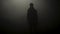 Black silhouette of man standing in smoky dark. Stock footage. Mysterious silhouette of young man stands in darkness lit