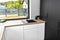 Black sink in a modern kitchen built into the top of kitchen cabinets against the wall with white fronts, visible corner window.