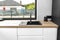 Black sink in a modern kitchen built into the top of kitchen cabinets against the wall with white fronts, visible corner window.