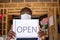 A black small business owner wearing a face mask and holding an open sign in front of his shop