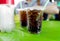 black sparkling cola water with bubbles in plastic cup with stroll on green table and empty plastic bottle beside blurred people