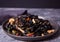 Black squid ink udon noodles with chicken meat on the black plate on dark stone background.