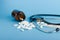 Black stethoscope, brown glass jar with white pills and thermometer on blue background. Medical equipment for diagnosis