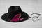 Black stylish hat with a pink flower lies next to fashionable eyewear on a white wooden table