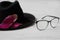 Black stylish hat with a pink flower lies next to fashionable eyewear on a white wooden table