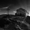 Black and white photo of small medieval Eastern Orthodox chapel and cross on top of a rocky hill. Sun rays backlight