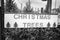 A black and white photograph of a painted white Christmas Tree sale sign hanging on a fence to a parking lot with evergreens