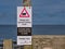 A black and white rectangular sign with a red warning triangle on a coastal path warns walkers