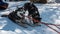 Black and white Siberian huskies are harnessed, resting on a snowy road.