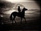 Black and white silhouette of a man riding a horse on a sandy beach under a cloudy sky during sunset