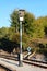 Black and white tall metal pole with railway signal lights on top surrounded with gravel and railway tracks with dense trees and