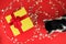 Black and white tuxedo cat with green eyes plays with golden gift box on bright red background with garland in form of