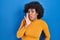 Black woman with curly hair standing over blue background hand on mouth telling secret rumor, whispering malicious talk