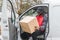 Black young adult delivery guy in work uniform sitting in door of white van looking at cardboard box parcel. Horizontal