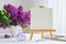 Blank canvas on easel, watercolor paints, brush for painting and lilac flowers on table